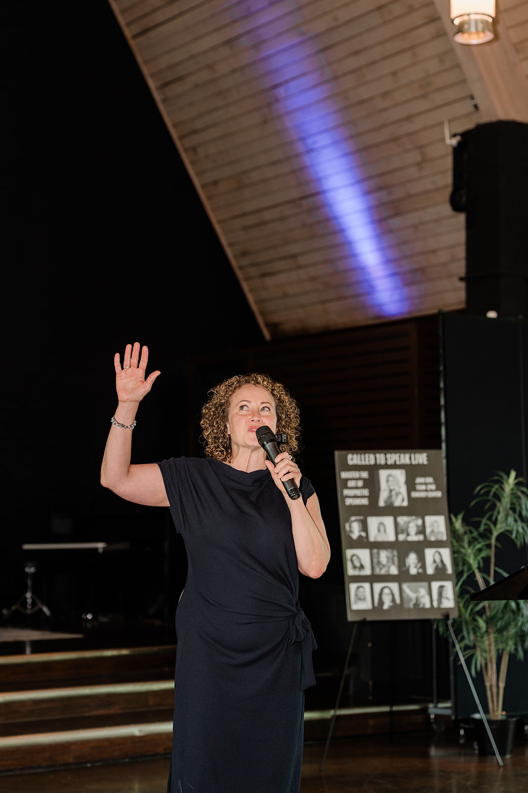 Woman with curly hair speaking into a microphone and raising her hand on stage, with a display board and plants nearby.