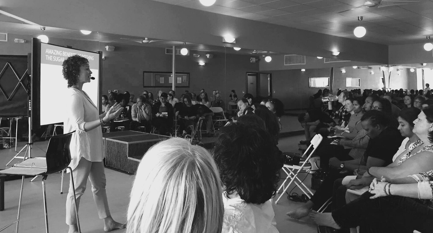 Woman presenting to a seated audience in a large room, with a screen behind her.