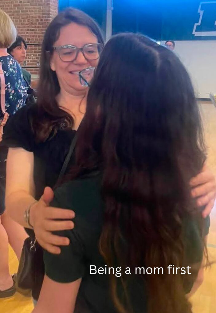 Two women hugging, one smiling and wearing glasses, the other with long dark hair, in a public setting.