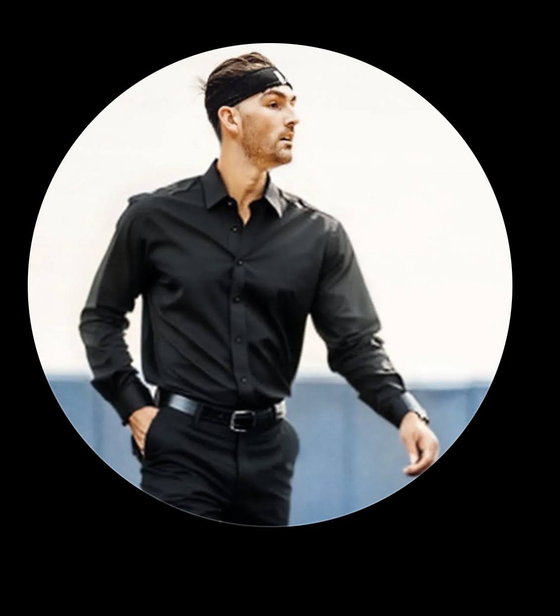 Man in black shirt and pants standing outdoors, looking to the side, with a blurred background.