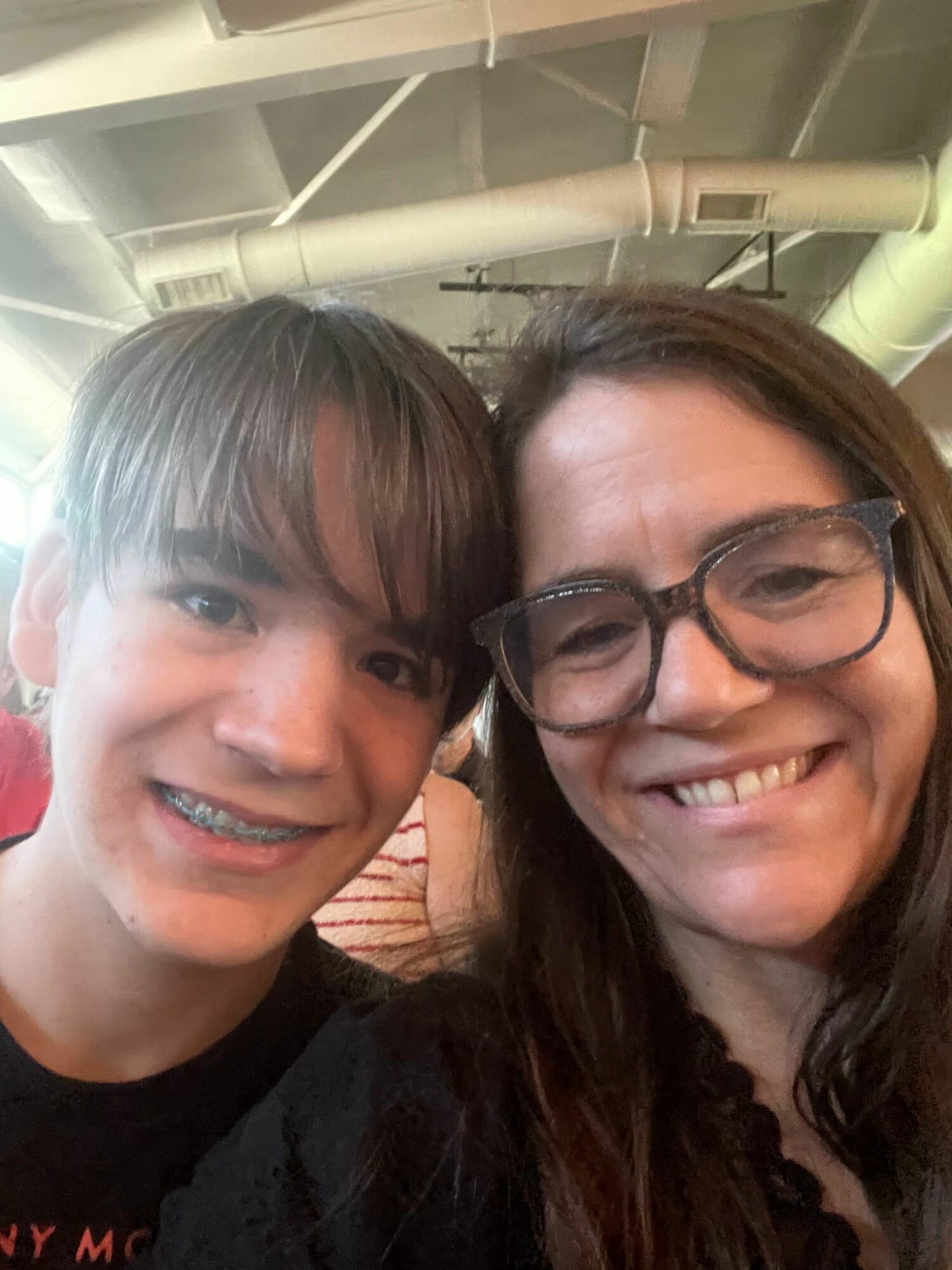 Two people smiling, one wearing glasses, close together indoors, with pipes visible in the background.