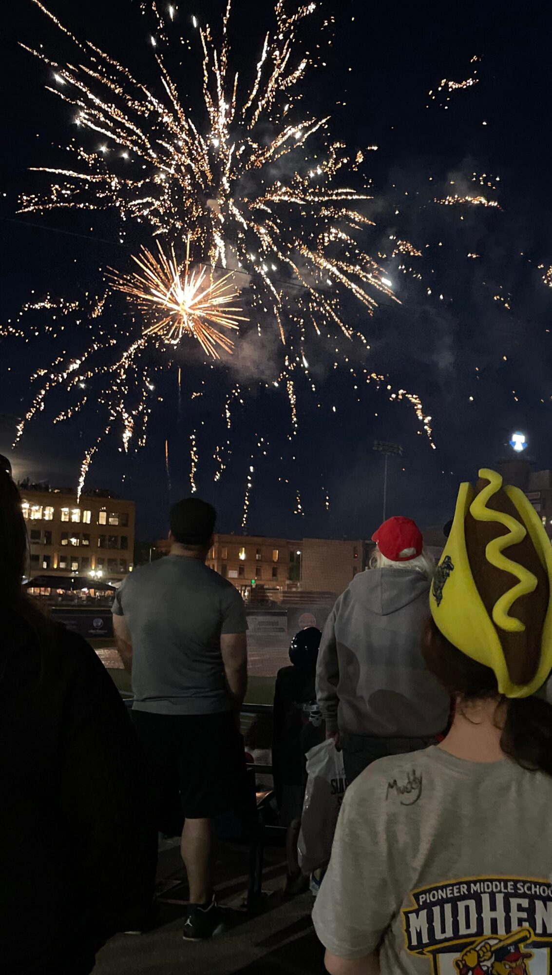 People watching fireworks display in night sky, some wearing hats, city buildings in background.