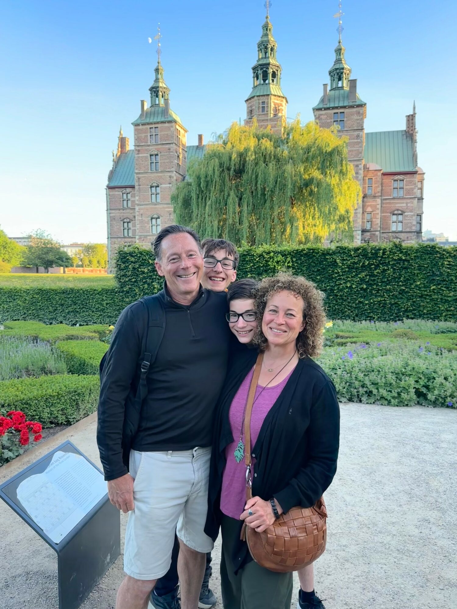 Four people standing outdoors in front of a historic castle with towers, green trees, and a hedge, smiling at the camera.