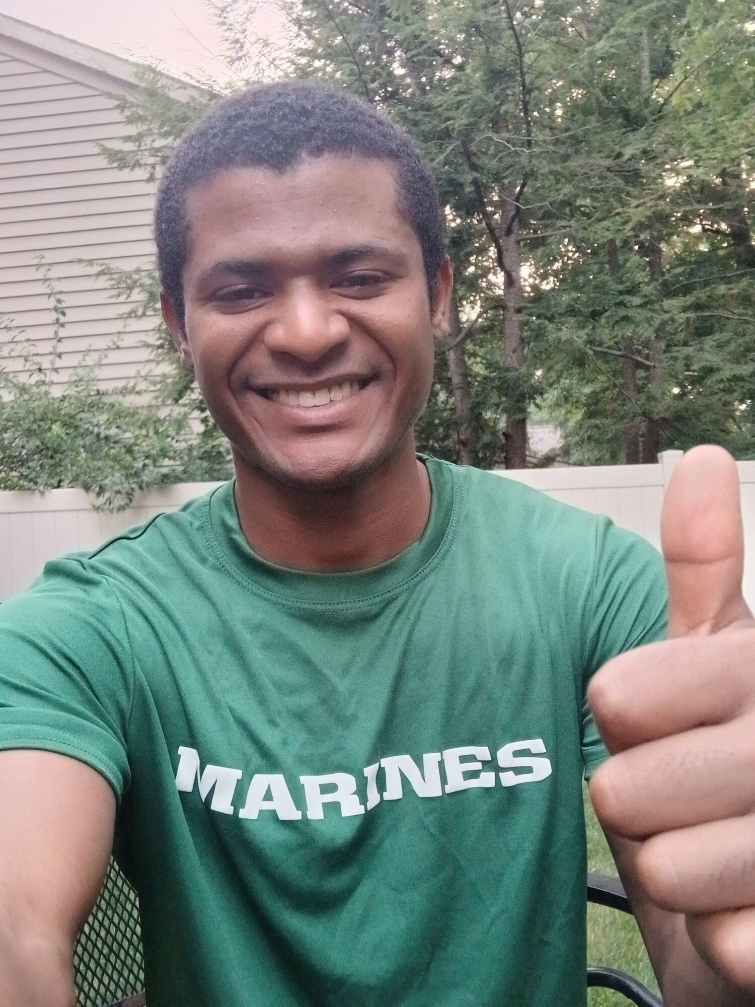 Young man smiling outdoors, wearing a green T-shirt with white text, sitting near a garden with trees and a house in the background.
