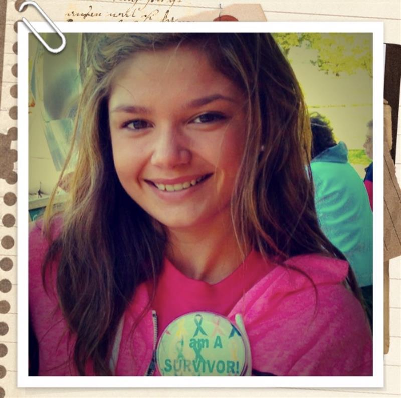 Smiling young woman with long hair wearing a pink hoodie, outdoors with trees and people in background.