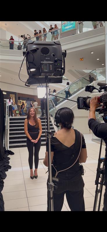 A woman stands in front of a camera setup in a shopping mall, with people on the upper level observing.