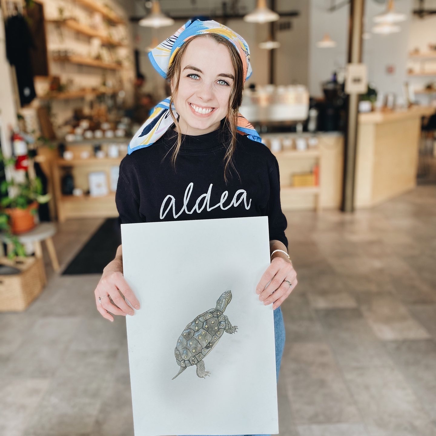 Smiling woman holding a drawing of a turtle in a store, wearing a black sweater and a colorful headscarf.
