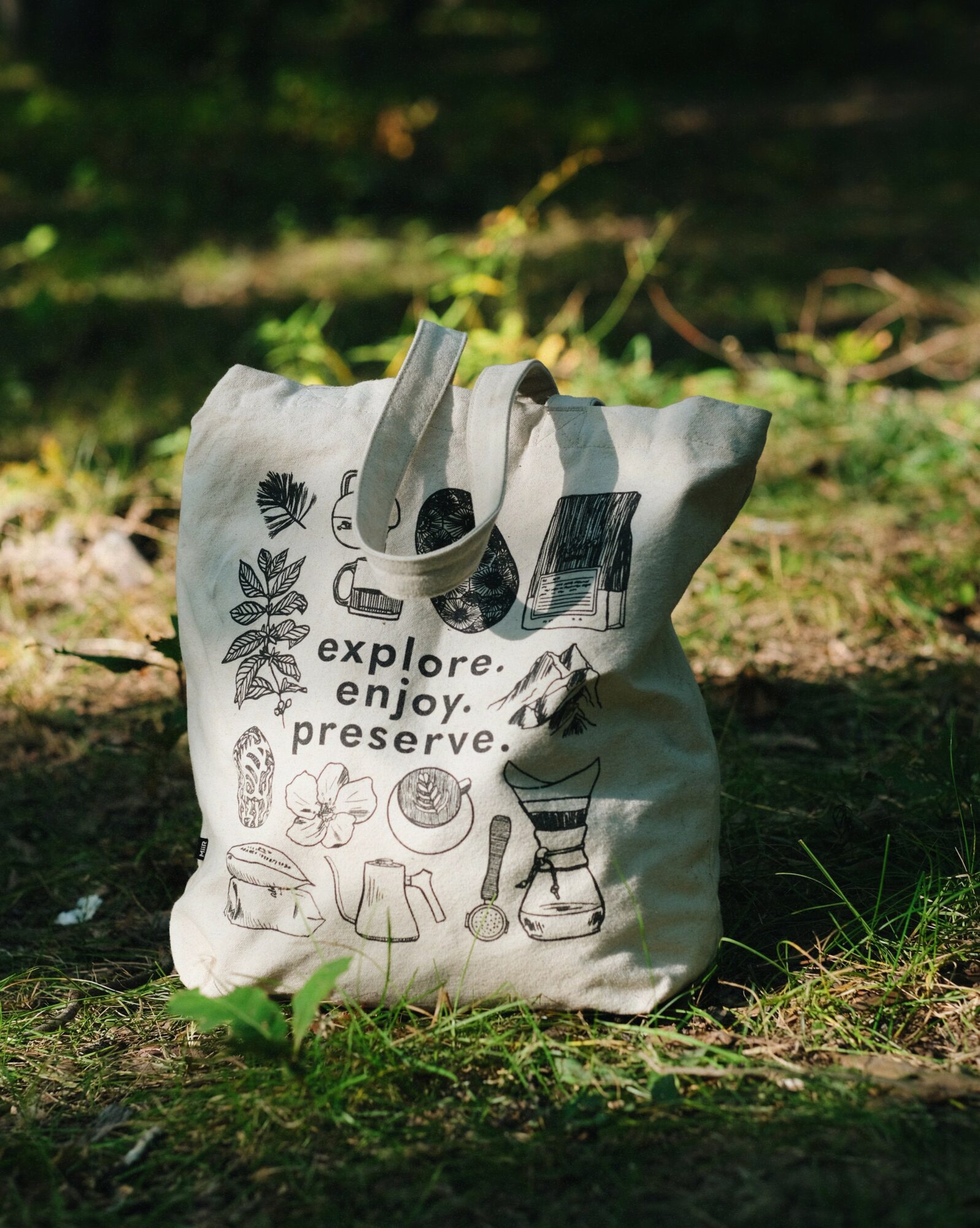 A white tote bag with black illustrations and text, placed on grass in sunlight, outdoors.