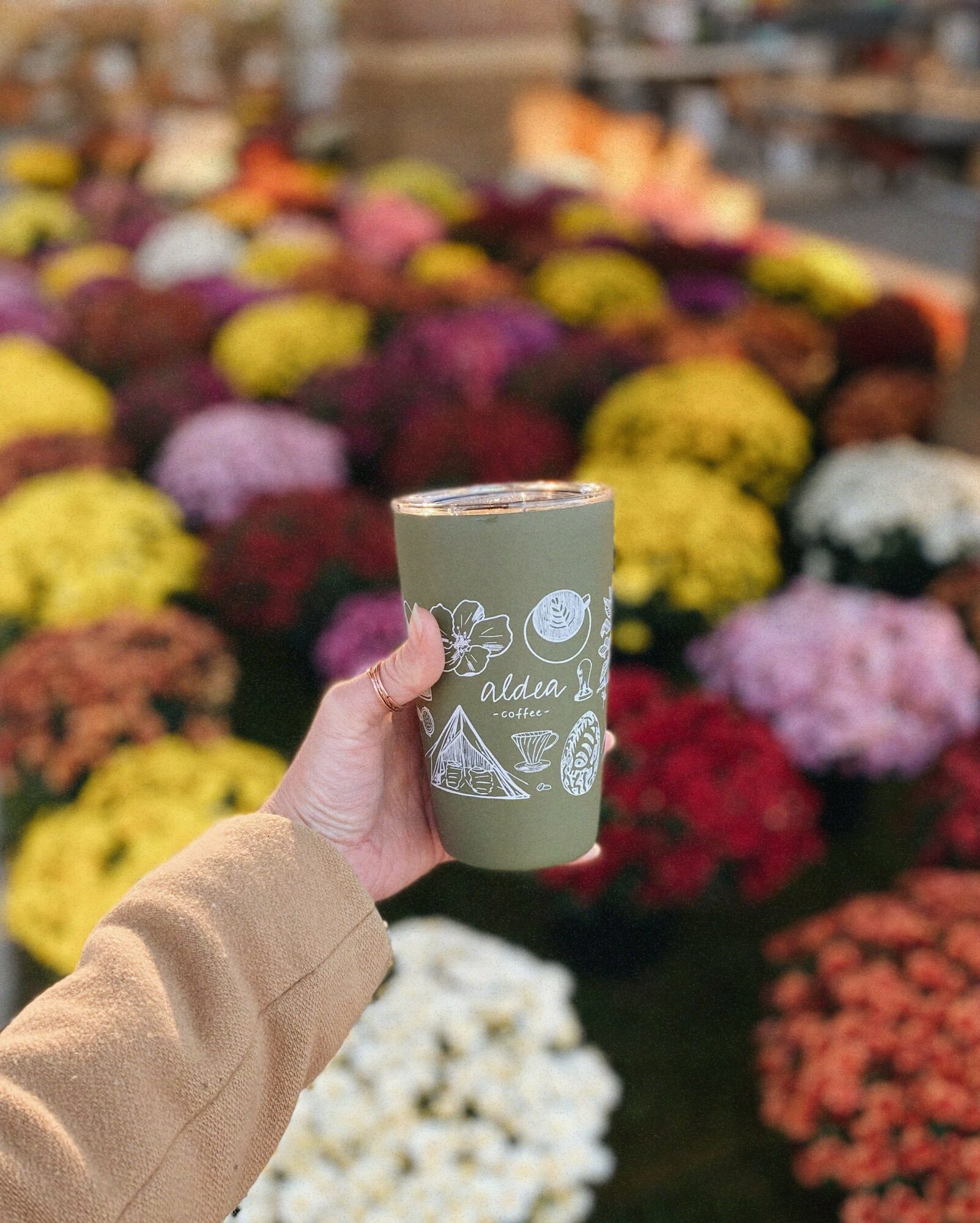 Person holding a green beverage in a clear cup with floral and fruit illustrations, in front of colorful flowers.