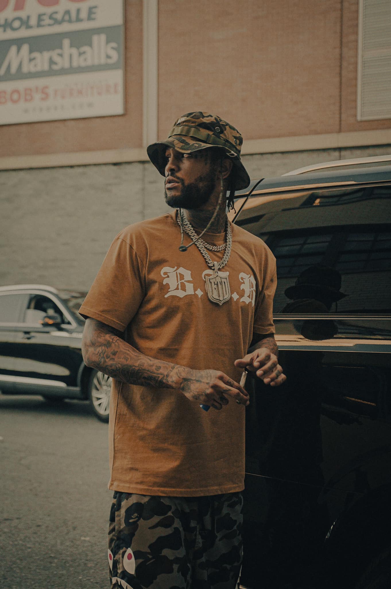 Man with tattoos, wearing a camouflage hat, gold chains, and a brown T-shirt, standing next to a black vehicle on street.