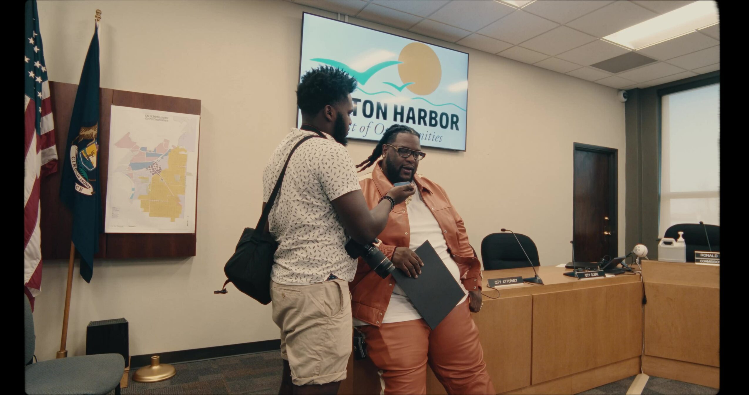 Two people talking in a meeting room with a city map and a screen displaying 'Benton Harbor'.