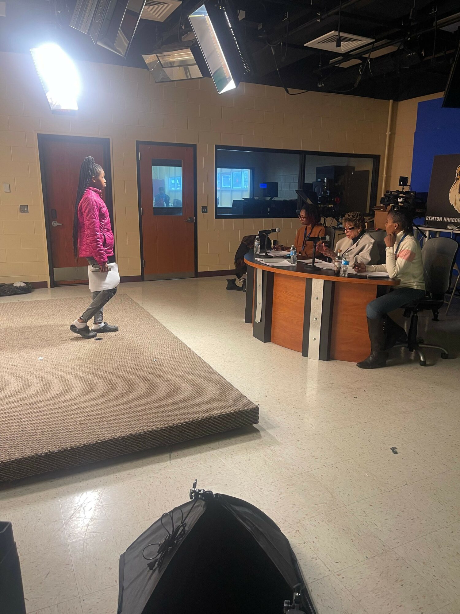 Person in pink jacket walking in an office with a large screen and desks, carpeted floor, and a black bag nearby.