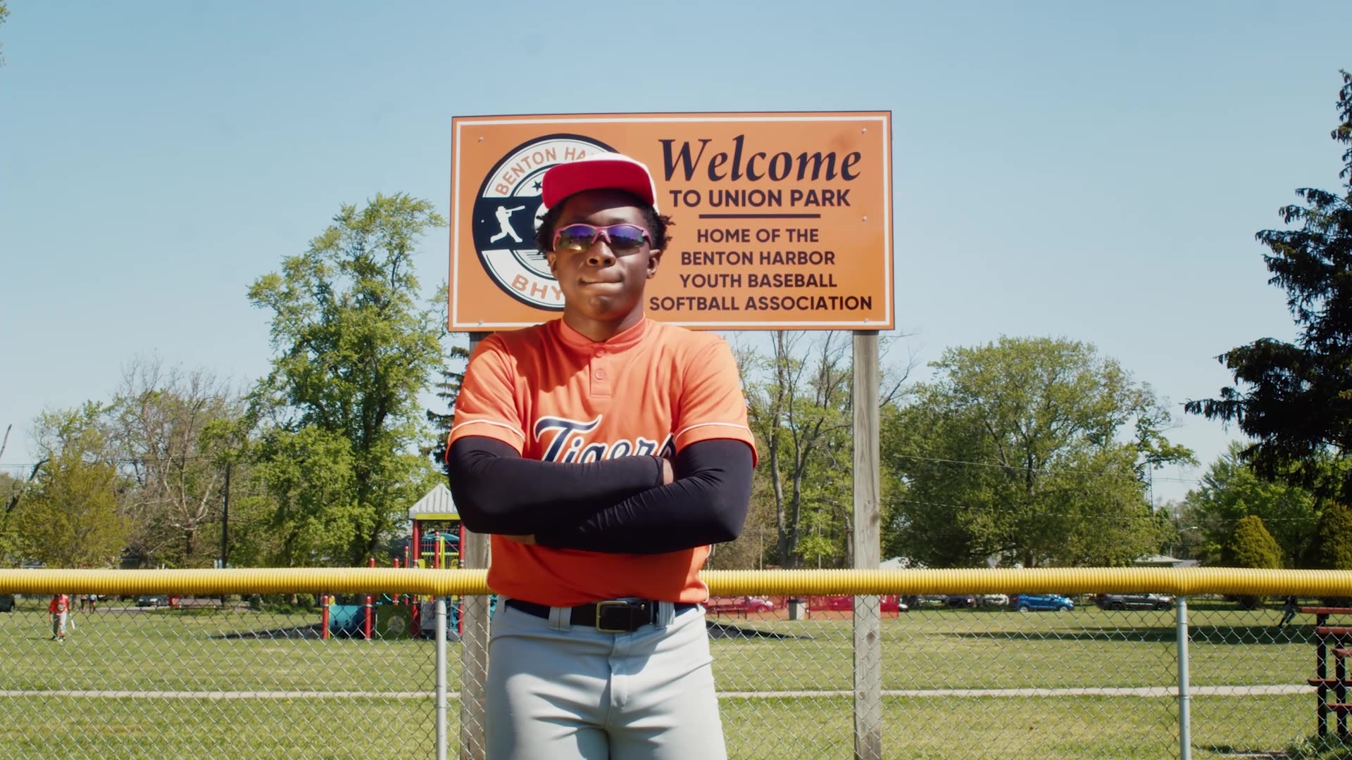 Person wearing sunglasses, red cap, orange shirt, and gray pants standing with arms crossed in front of a sign at a baseball field.