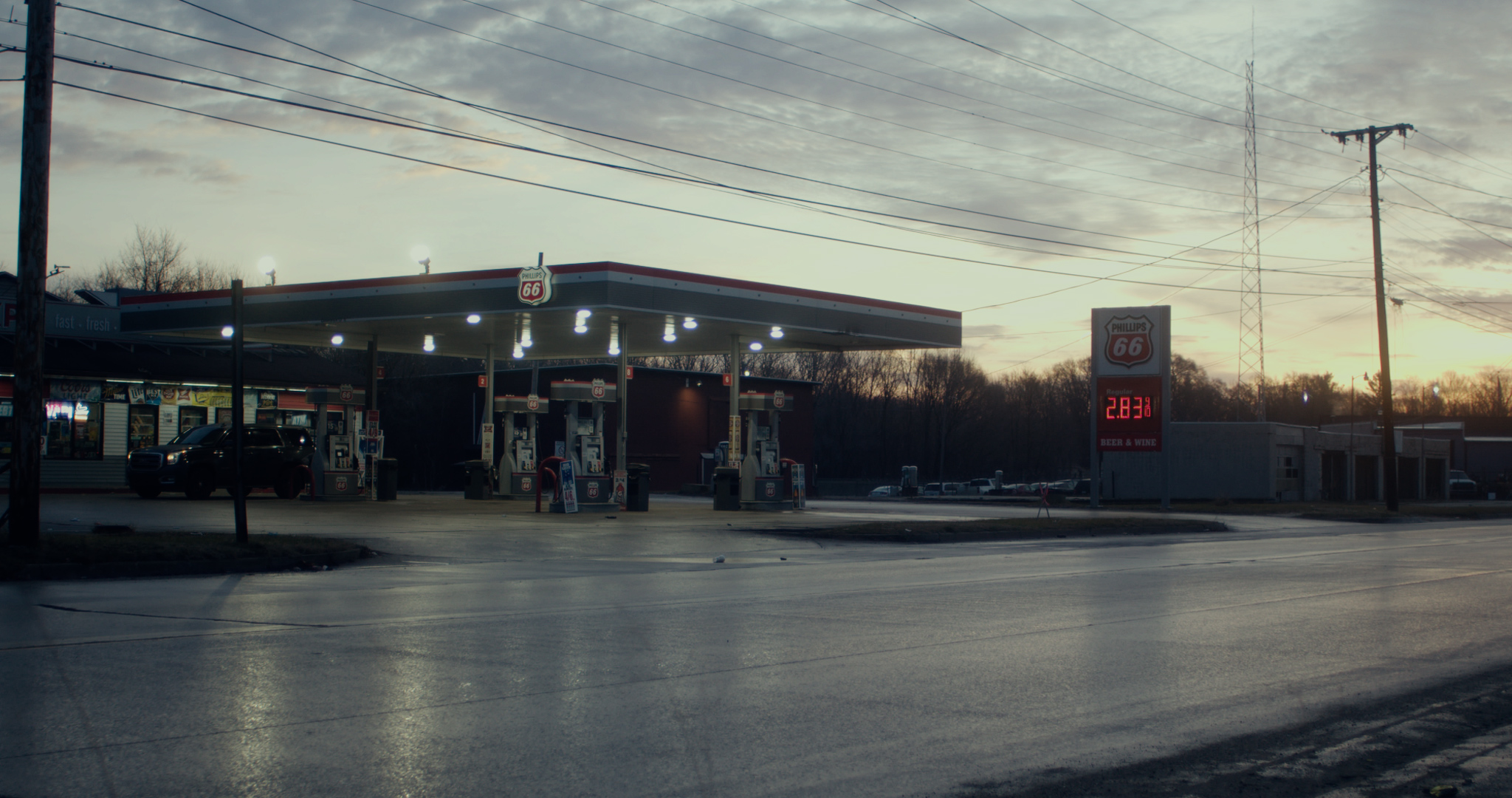 Gas station with fuel pumps, canopy, and a digital price sign, under a cloudy sky during sunset.