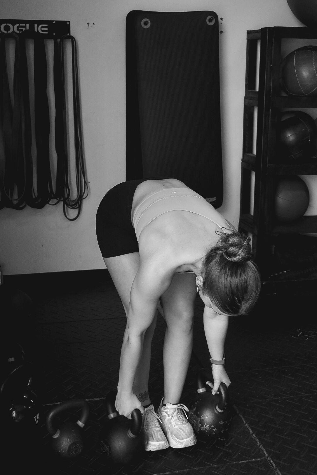 Person bending forward to pick up kettlebell in a gym, with equipment on wall and shelves nearby.