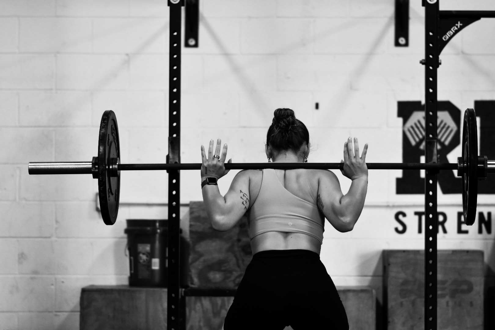 Woman lifting a barbell in a gym, facing a wall with gym equipment and a logo, black and white image.