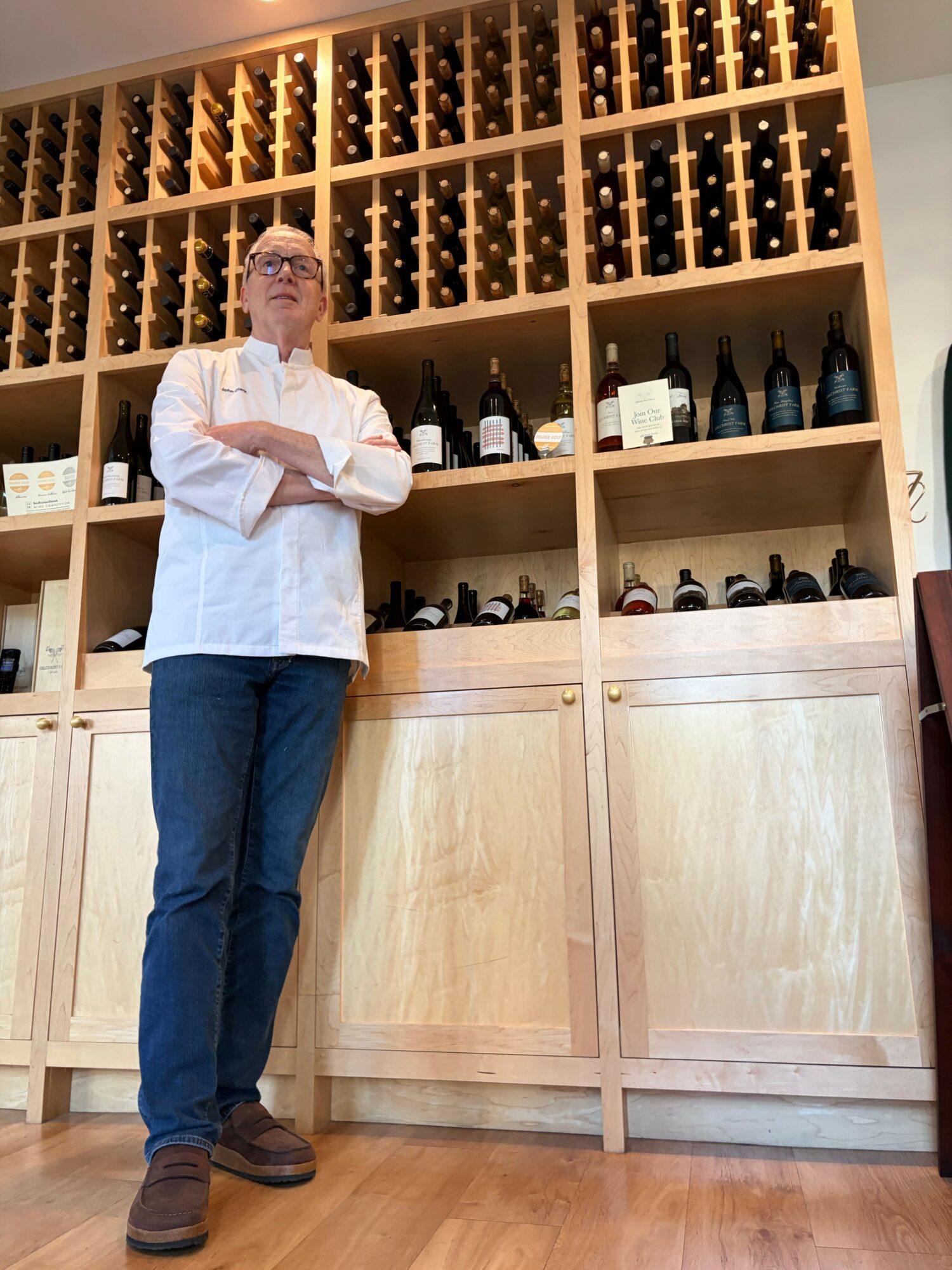 Person standing in front of wine racks, arms crossed, wearing a white shirt and blue jeans, in a wine cellar.