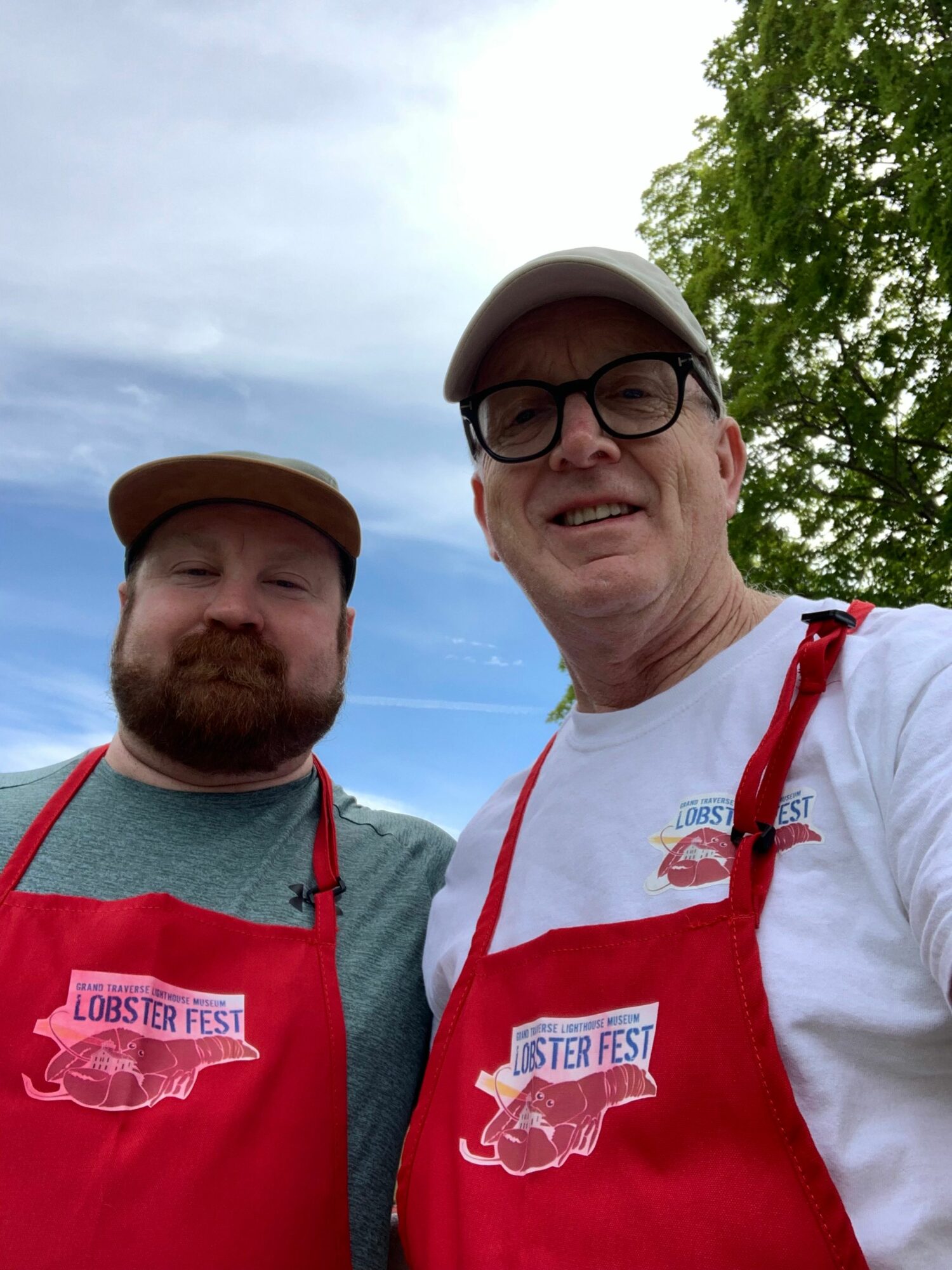 Two men outdoors, one with glasses and a cap, the other with a brown hat, both wearing jackets with logos, smiling.