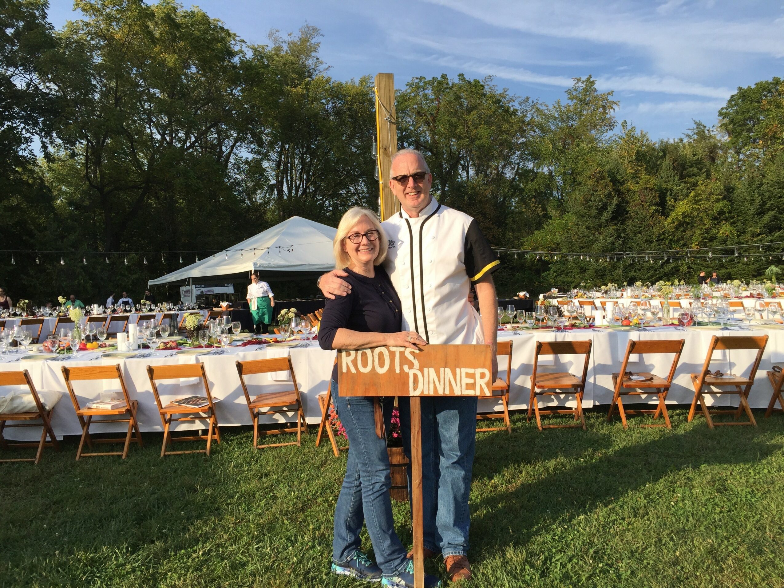 Two people standing outdoors near a long table with chairs, with trees and a tent in the background.
