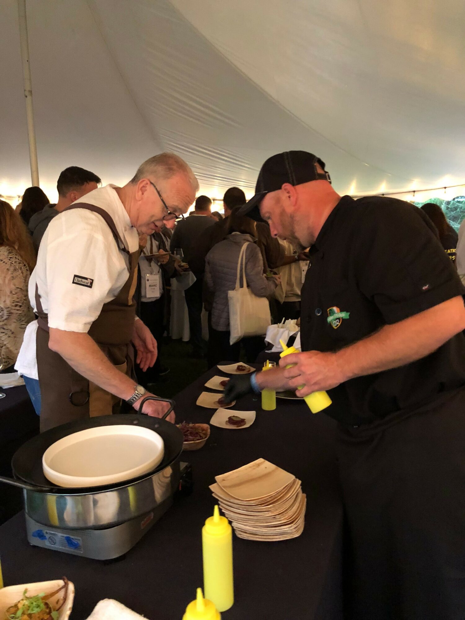 People gathered around a table at an indoor event, with one person serving food or drinks, others waiting in line.