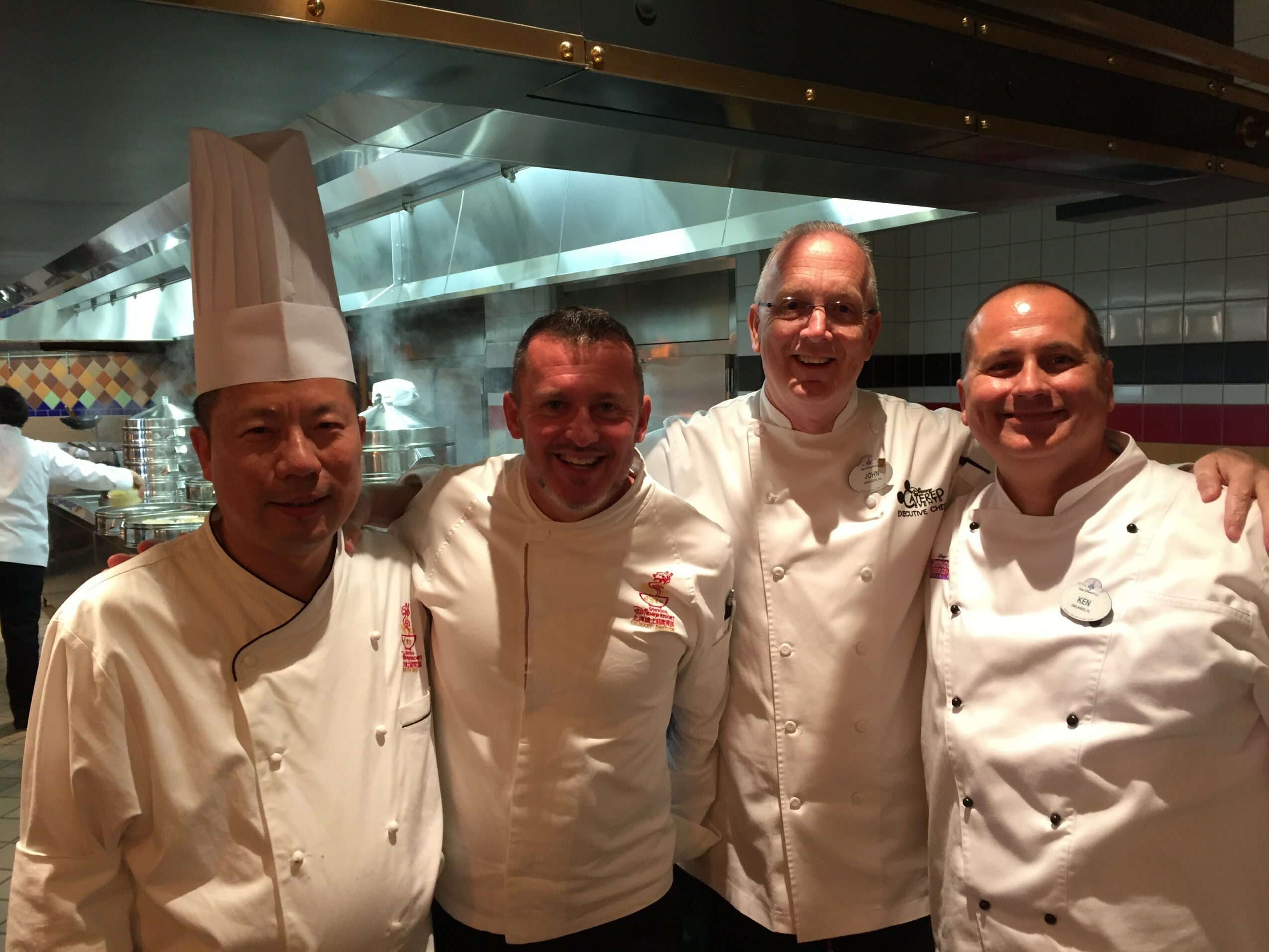 Four chefs in white uniforms standing together in a kitchen, smiling, with a stainless steel background.