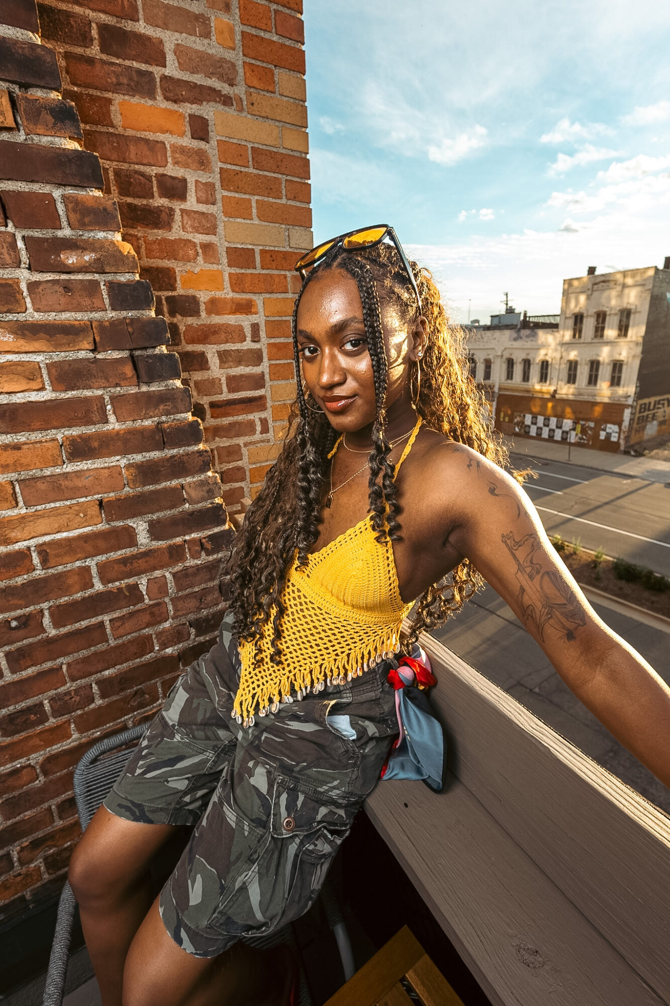 Young woman with long curly hair wearing a yellow top and camouflage shorts, leaning against a brick wall outdoors.