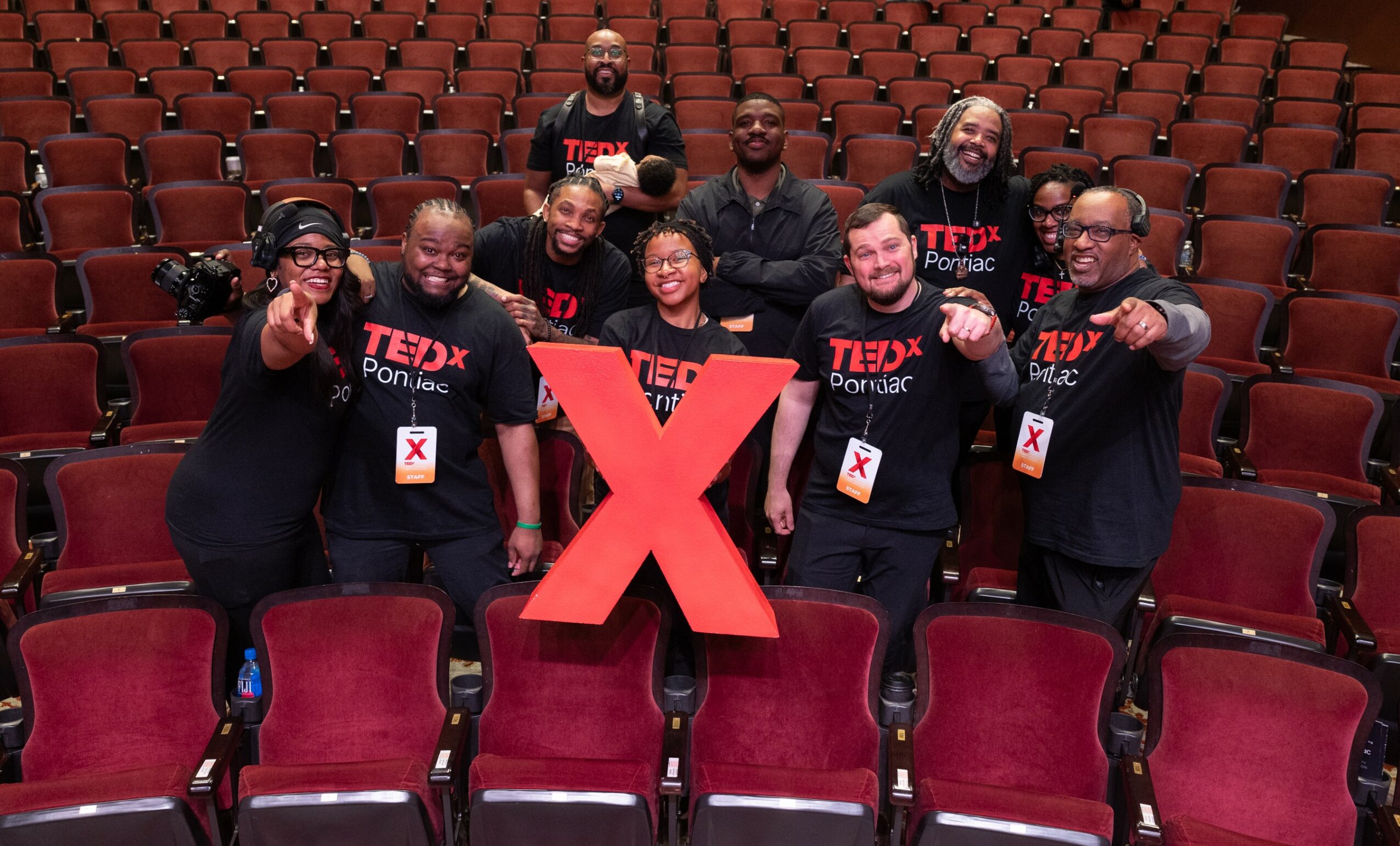 Group of nine people standing in an auditorium with red seats, holding a large red X, wearing matching T-shirts.