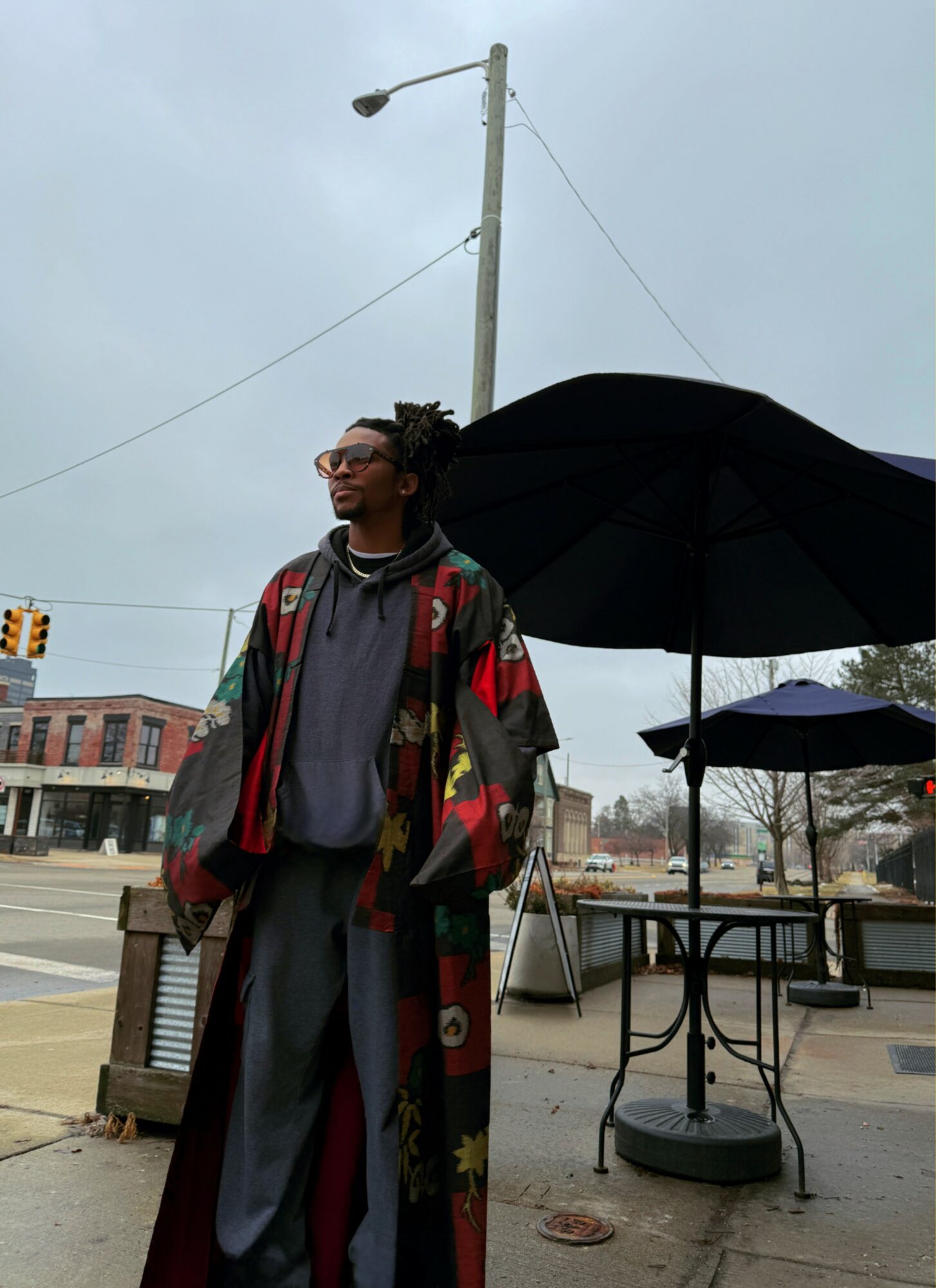 Person standing outdoors under an umbrella, wearing a colorful jacket and glasses, with a streetlight and buildings in the background.