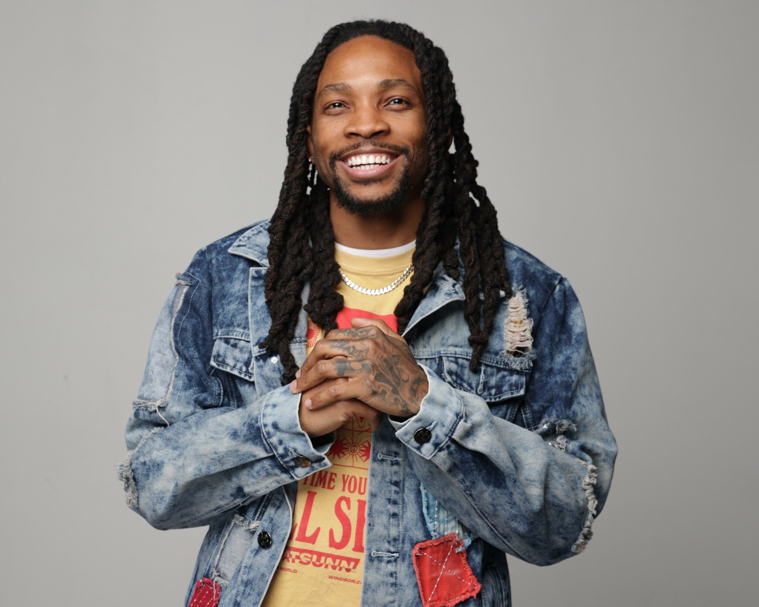 Smiling person with long dreadlocks wearing a denim jacket and layered necklaces, holding hands together, against a plain background.