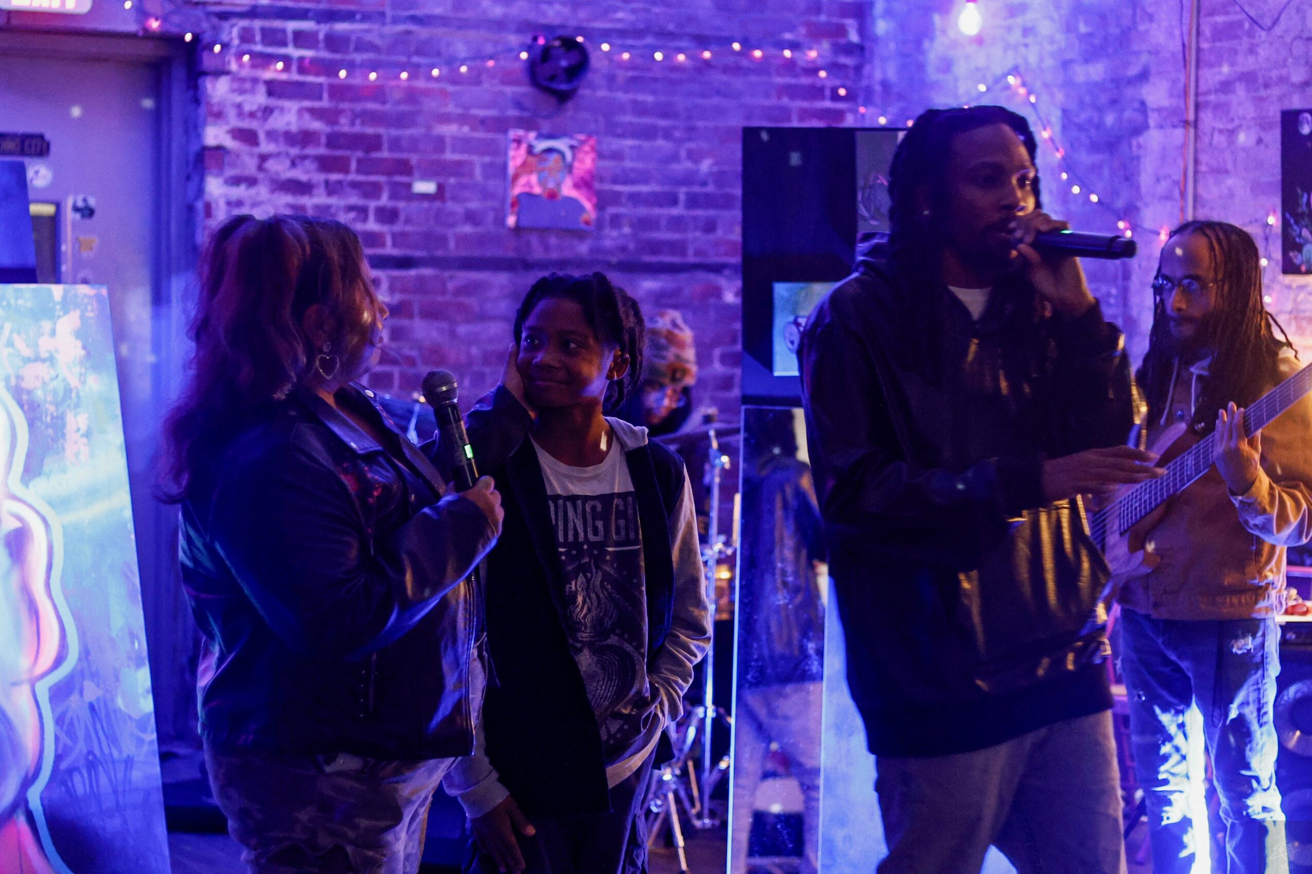 Group of four people in a dimly lit venue with purple lighting, one person speaking into a microphone, others listening.
