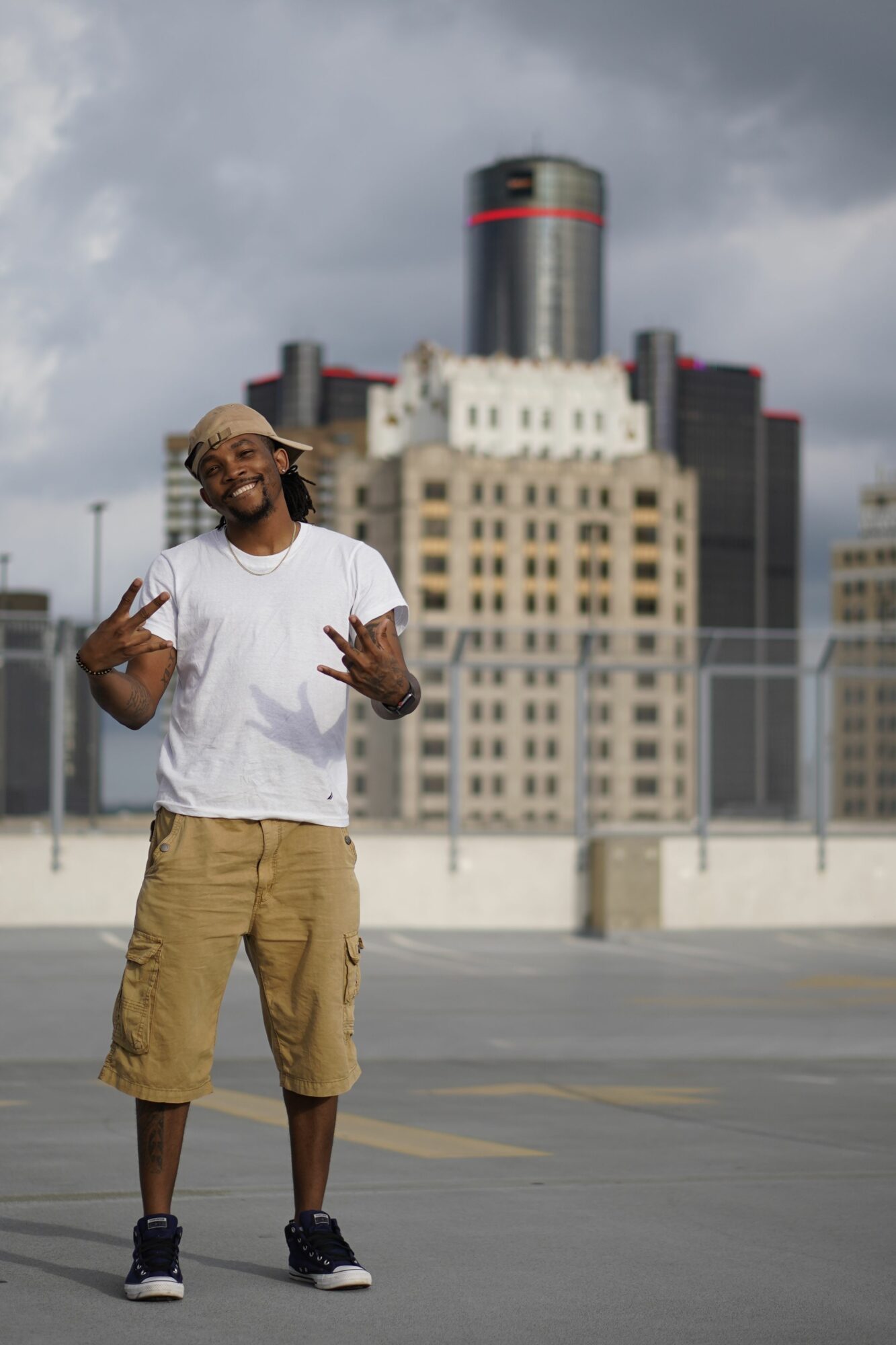 Person standing outdoors in an urban area with tall buildings, wearing a white shirt and tan pants, holding a phone.