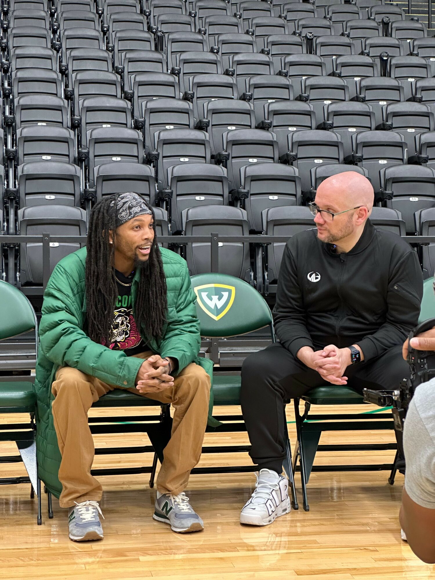 Two people sitting on chairs in an indoor setting with empty seats in the background, one wearing a black jacket and the other a green jacket.