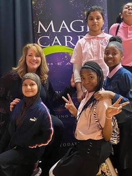 Group of five people, including a woman and four girls, posing in front of a purple and green banner. One girl makes a peace sign.