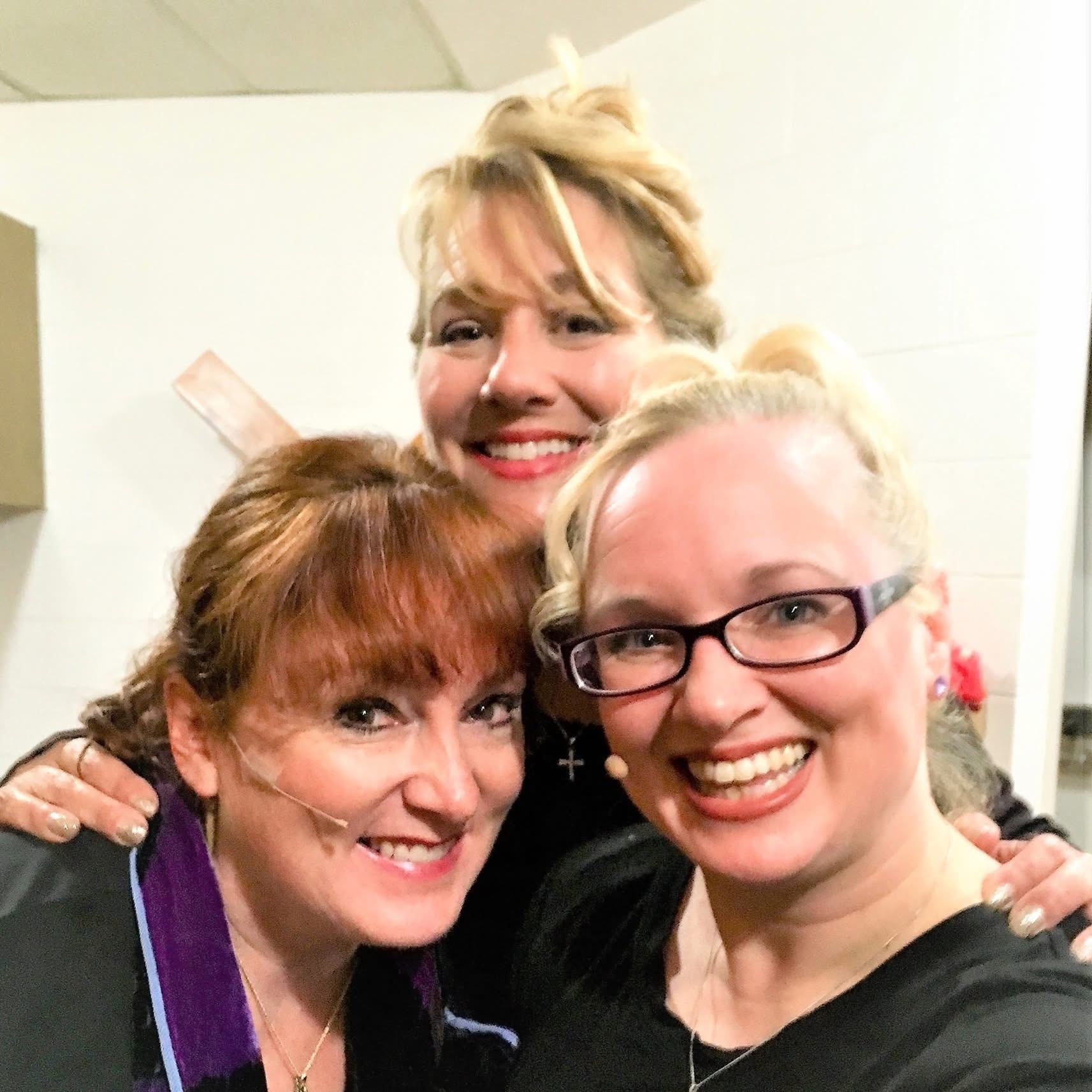 Three women smiling closely together indoors, one with glasses, all with light-colored hair.