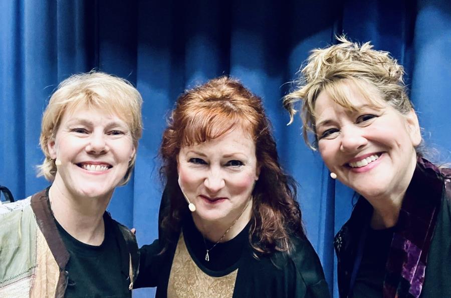 Three women smiling in front of a blue curtain, close together, facing the camera.
