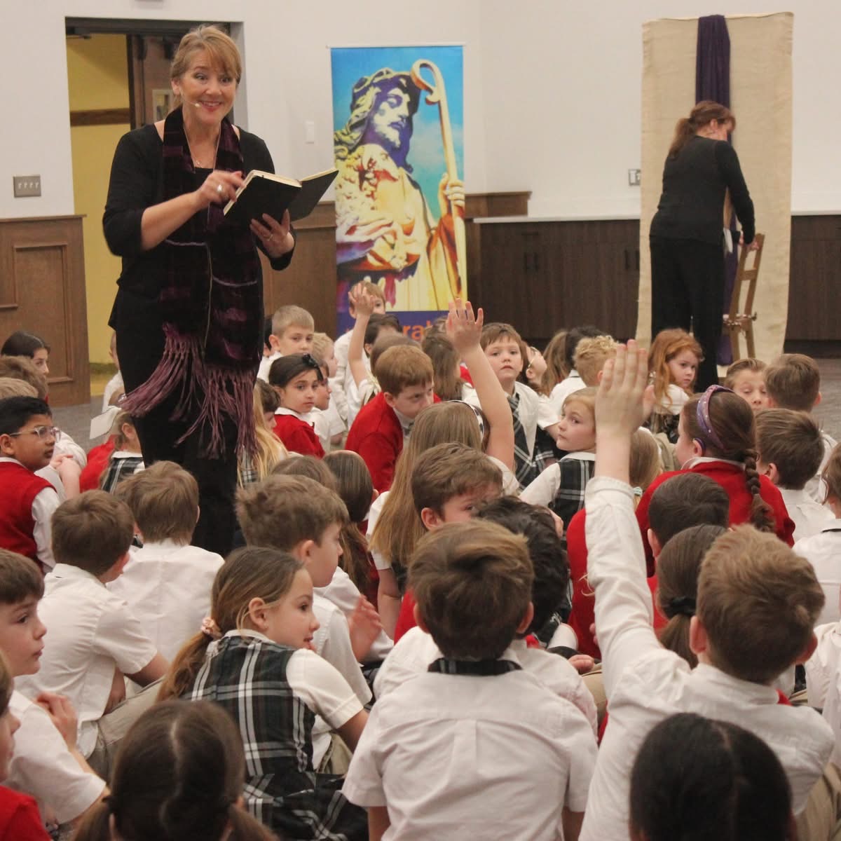 Woman standing with a book in front of children, some children raising hands, classroom setting with artwork in background.