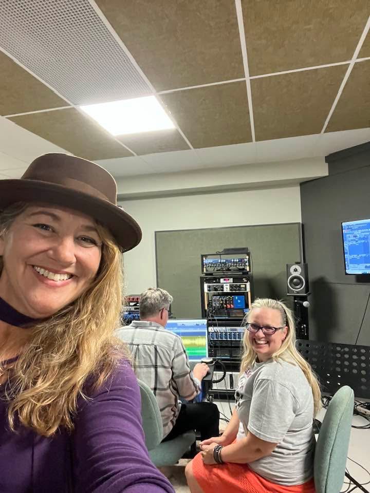 Two women and a man in a recording studio with audio equipment, smiling at the camera, woman in foreground wearing a hat.