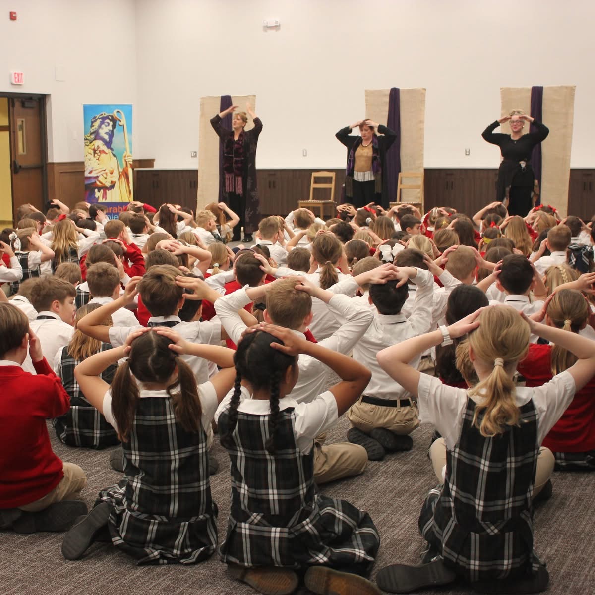 Children sitting on the floor watching a performance with four women holding boxes on their heads, in a school auditorium.