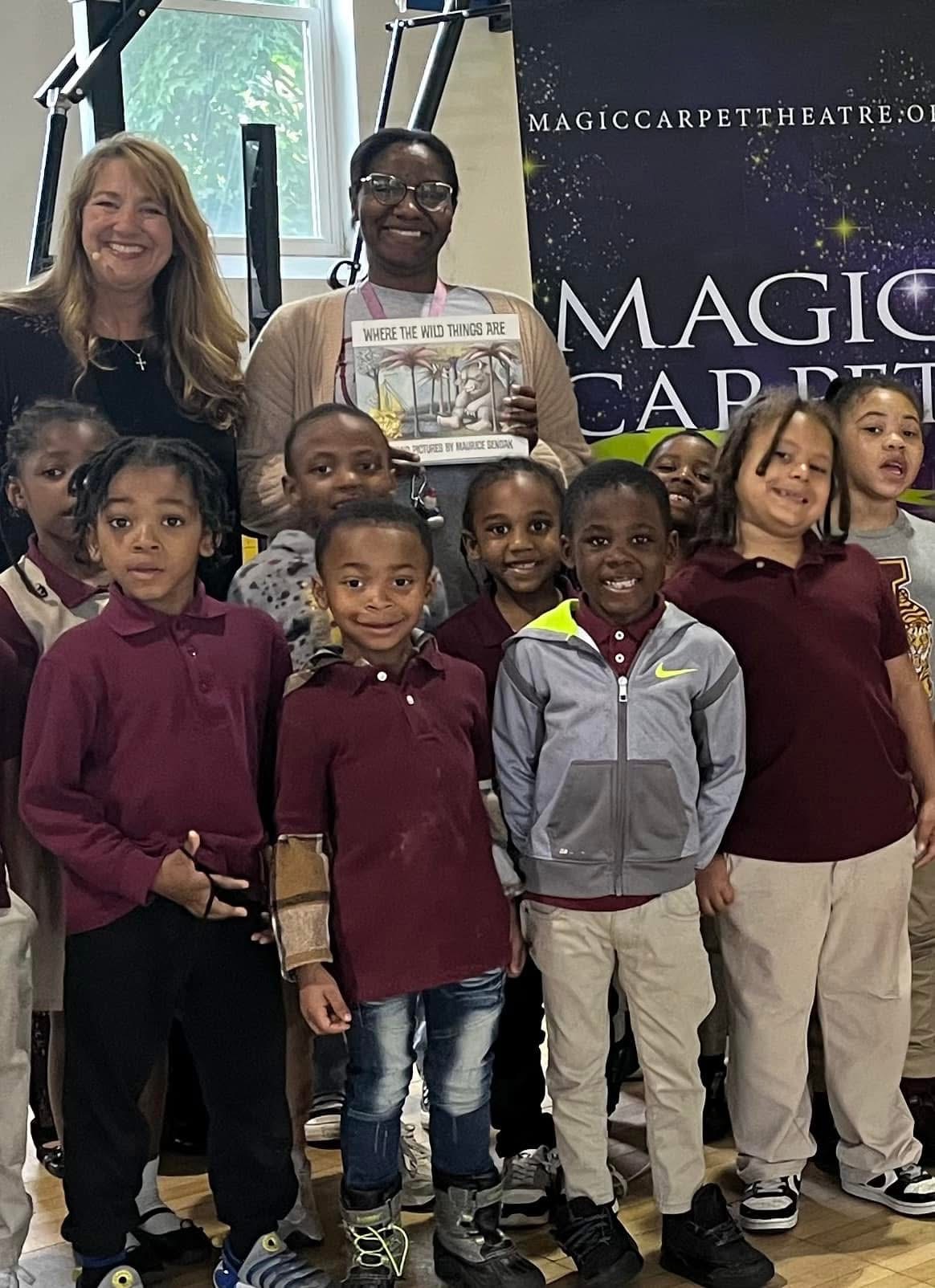 Group of children and two adults standing indoors, smiling, with a banner reading 'Magic Carpet Theatre' in the background.