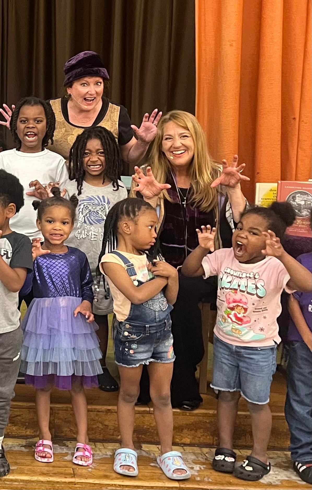 Group of children and two women smiling and waving in front of colorful curtains, some children wearing dresses and casual clothes.