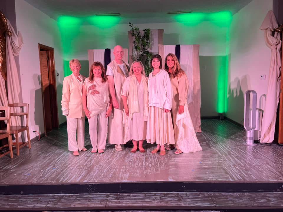 Group of seven people standing on stage with green lighting, some wearing white or light-colored clothing, in a room with curtains and chairs