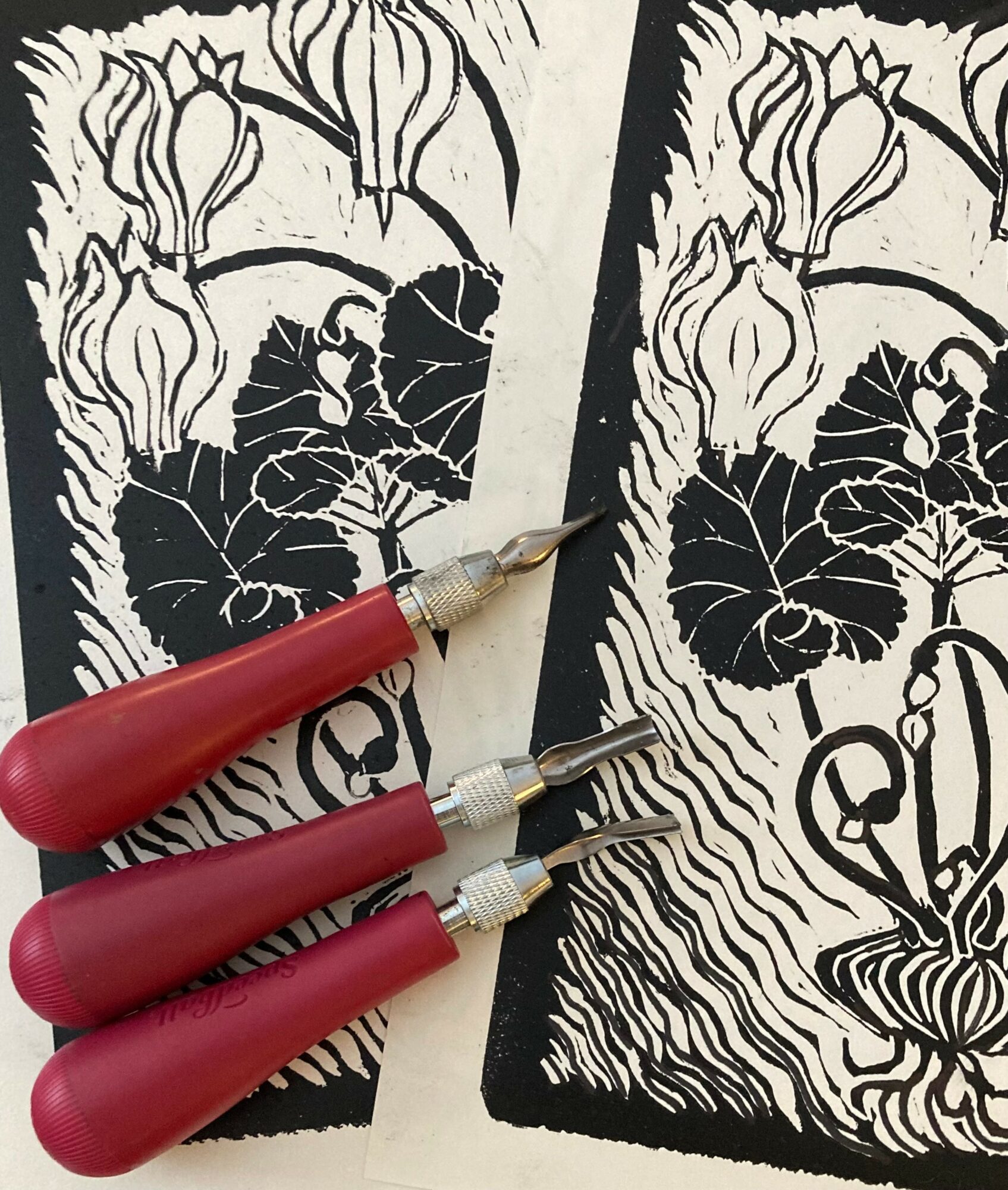 Two black and white paper cutouts of flowers and leaves, with three red carving tools placed on top.