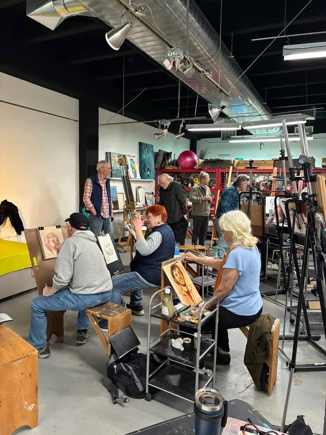 People sitting and painting at an art class in a studio with art supplies and artwork around.