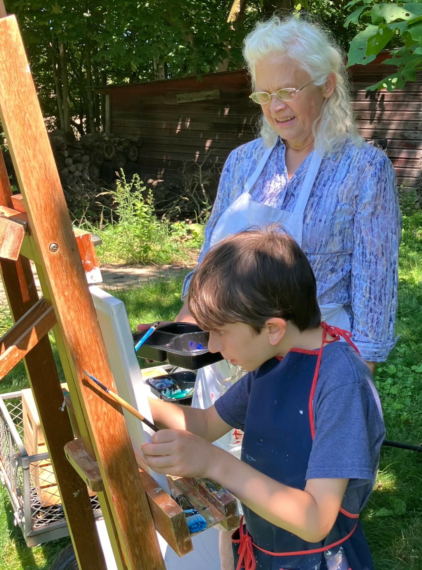 A young boy painting outdoors with an elderly woman watching, surrounded by greenery and a wooden fence.