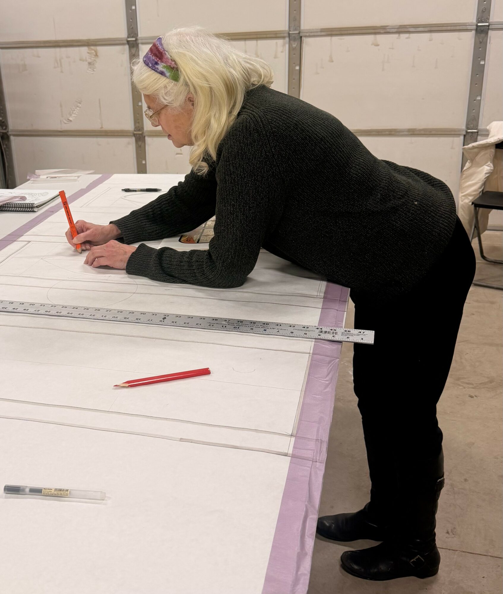 Woman with blonde hair, glasses, and a black sweater leans over a large table, writing with a red marker, in a garage-like space.