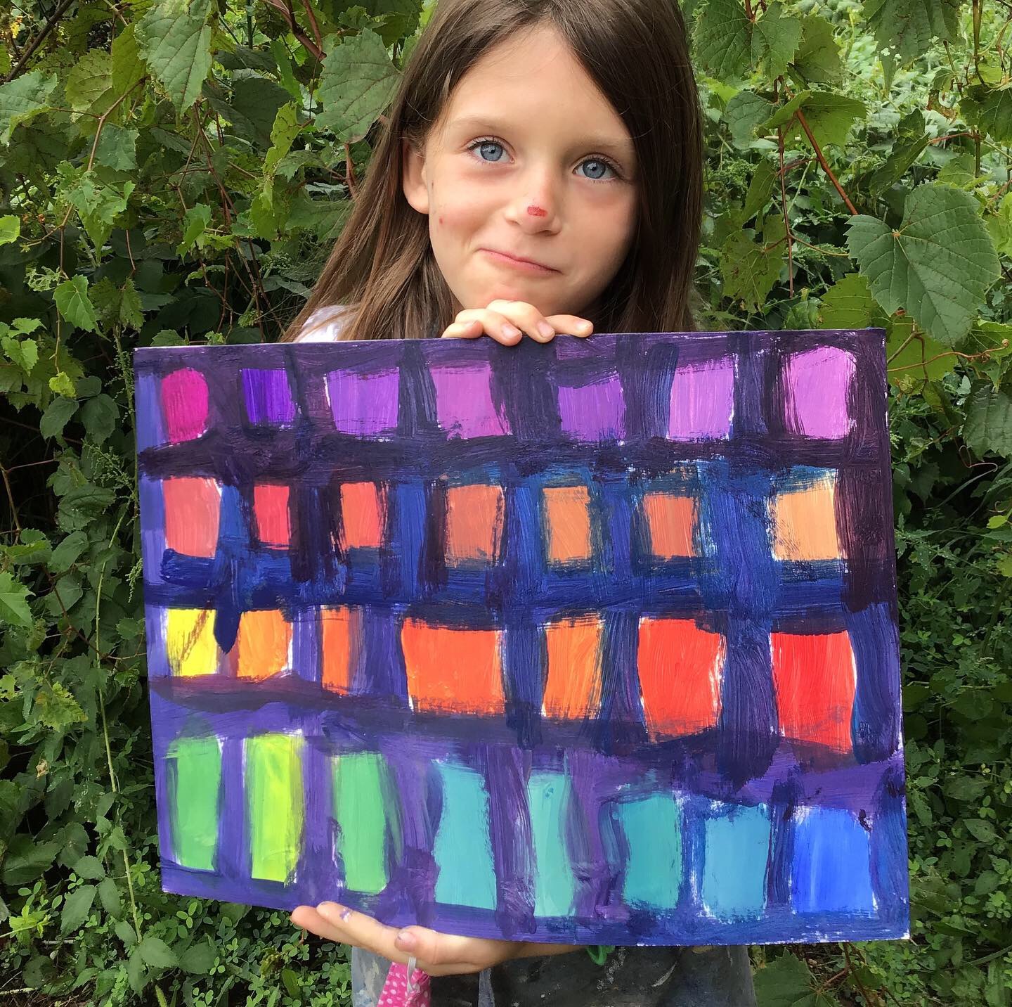 Young girl holding a colorful painted canvas outdoors with green foliage background.