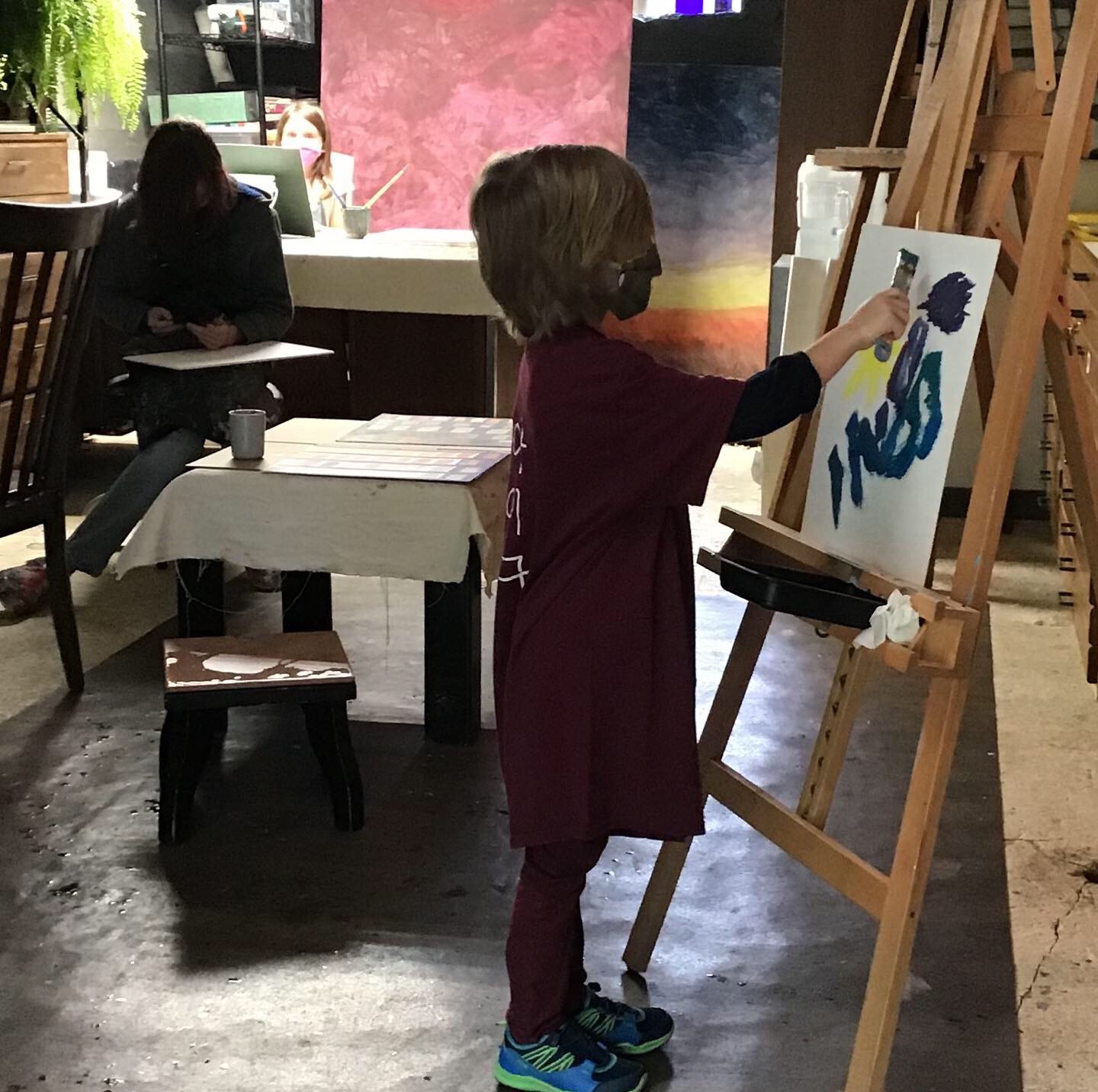 Child painting on an easel in a room with other people and art supplies, facing right, with a table nearby.