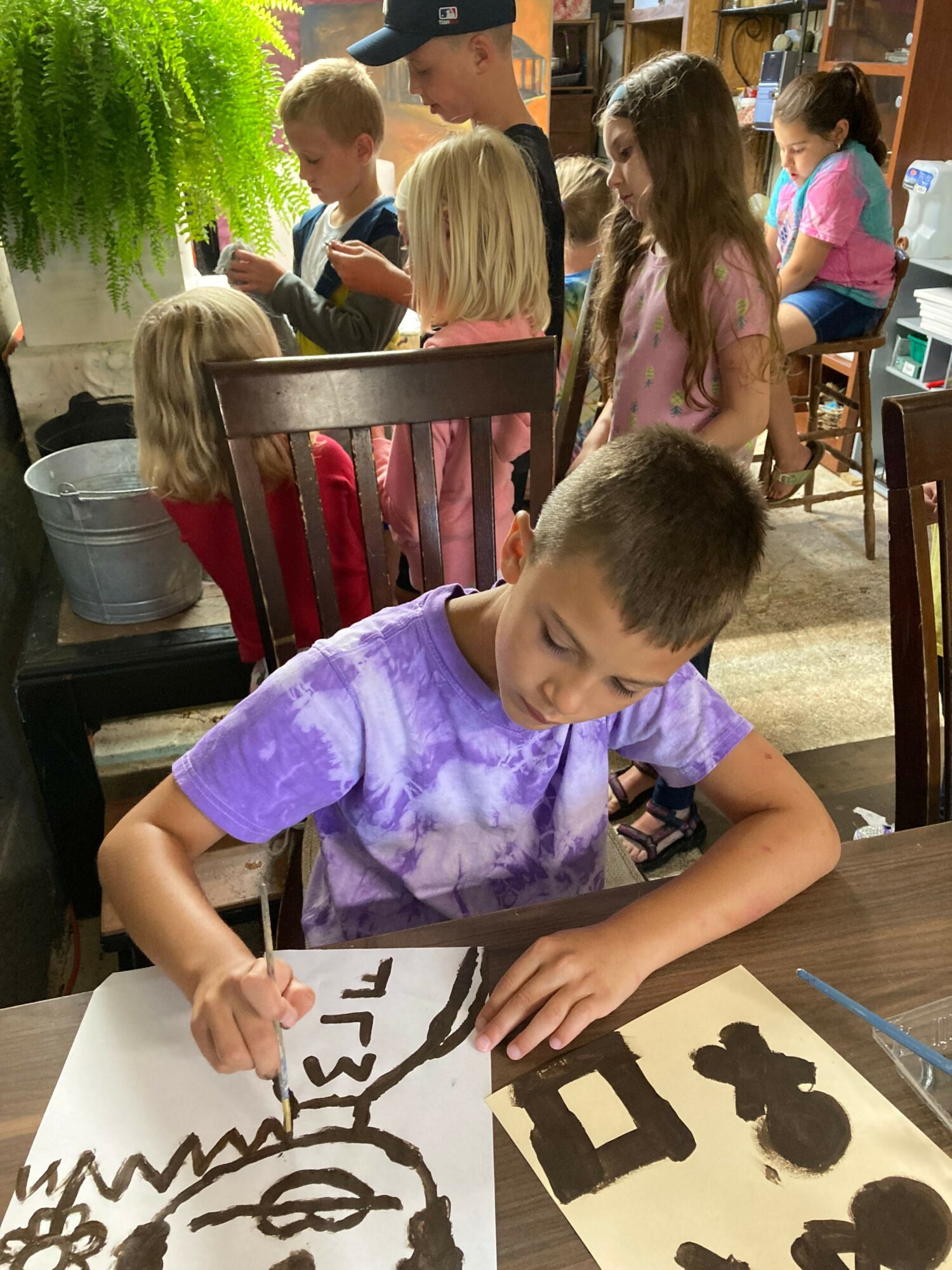 Child drawing on paper with black marker, surrounded by other children in a classroom setting.