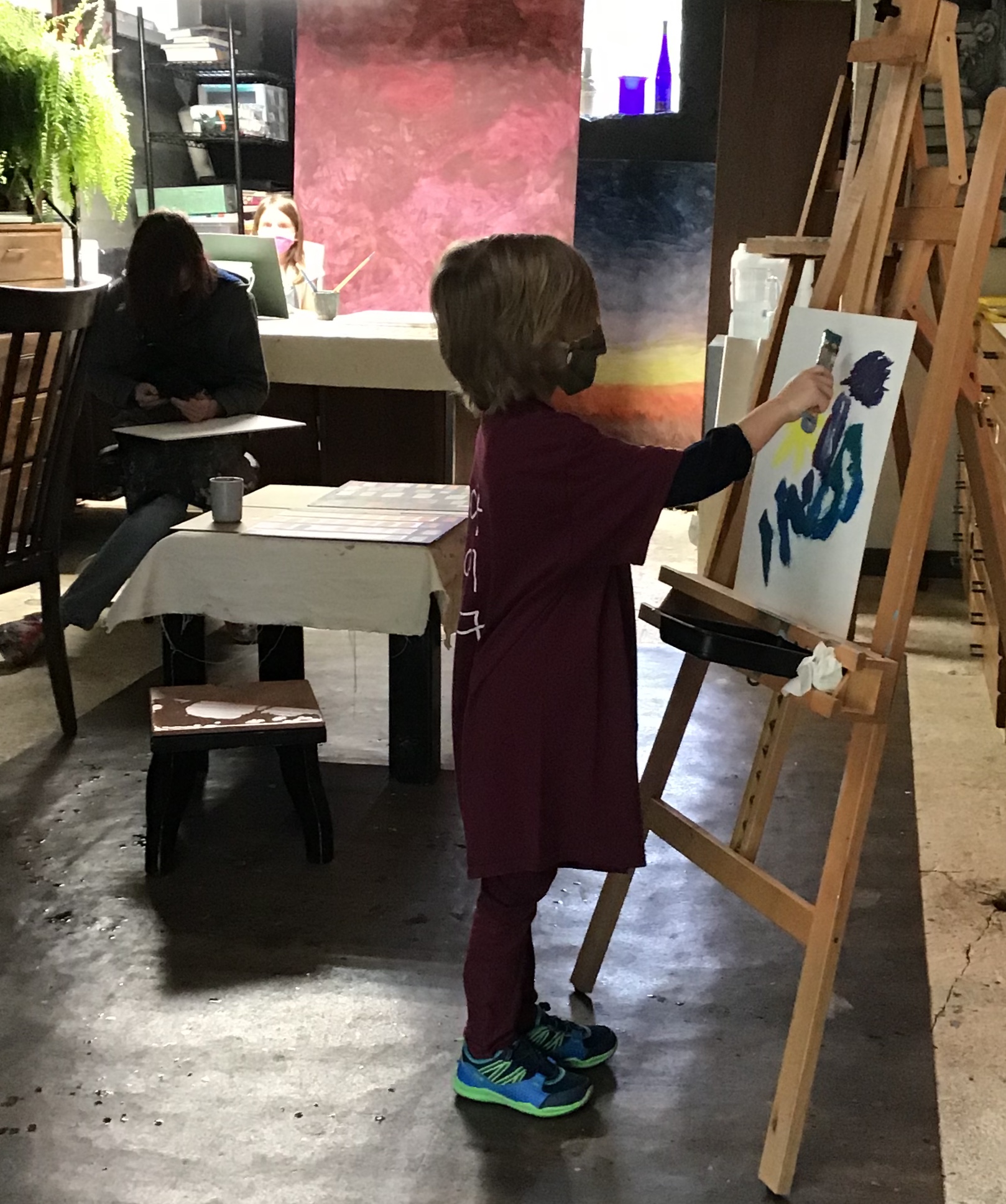 Child painting on canvas at an easel in an art studio, with a person sitting at a table in the background.
