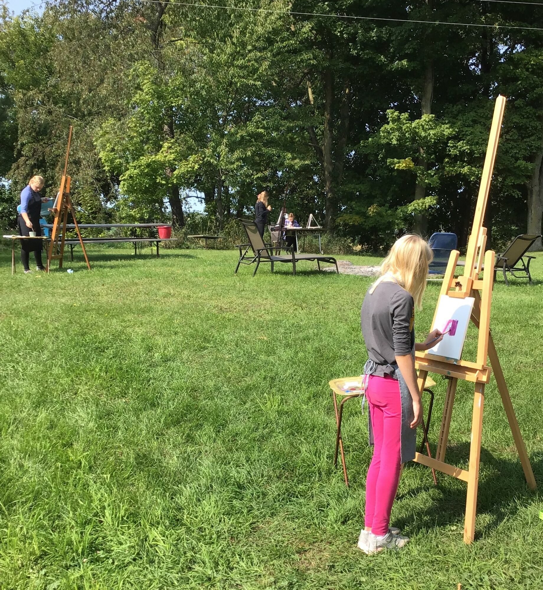 Person painting on an easel outdoors with trees and other people in the background.